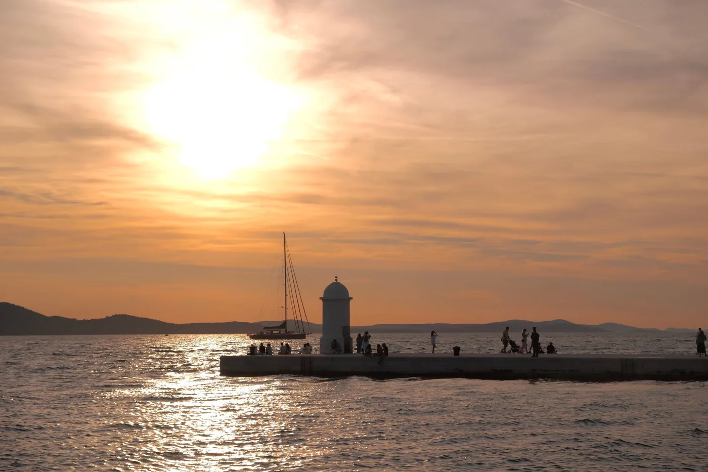 Eine steinerne Mole mit einem kleinen weißen Leuchtturm und einer Segeljacht im tiefen goldenen Licht