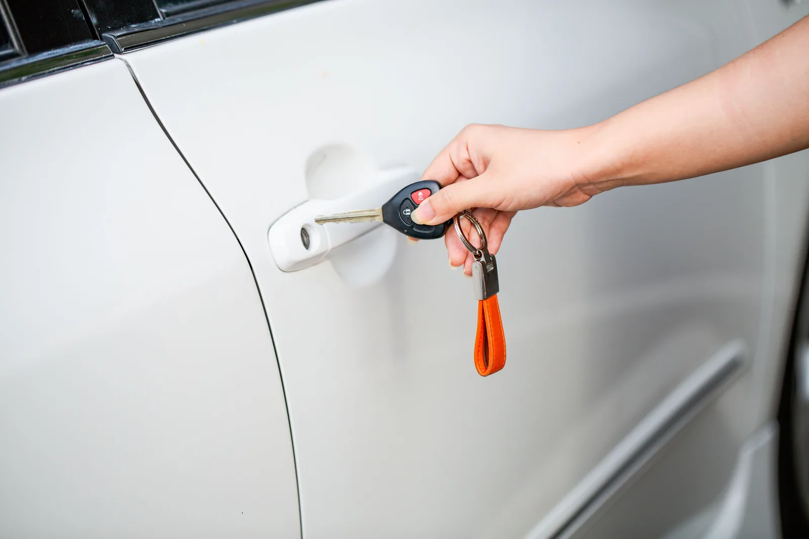 A hand holding a car key with a remote fob beside the door handle of a white car