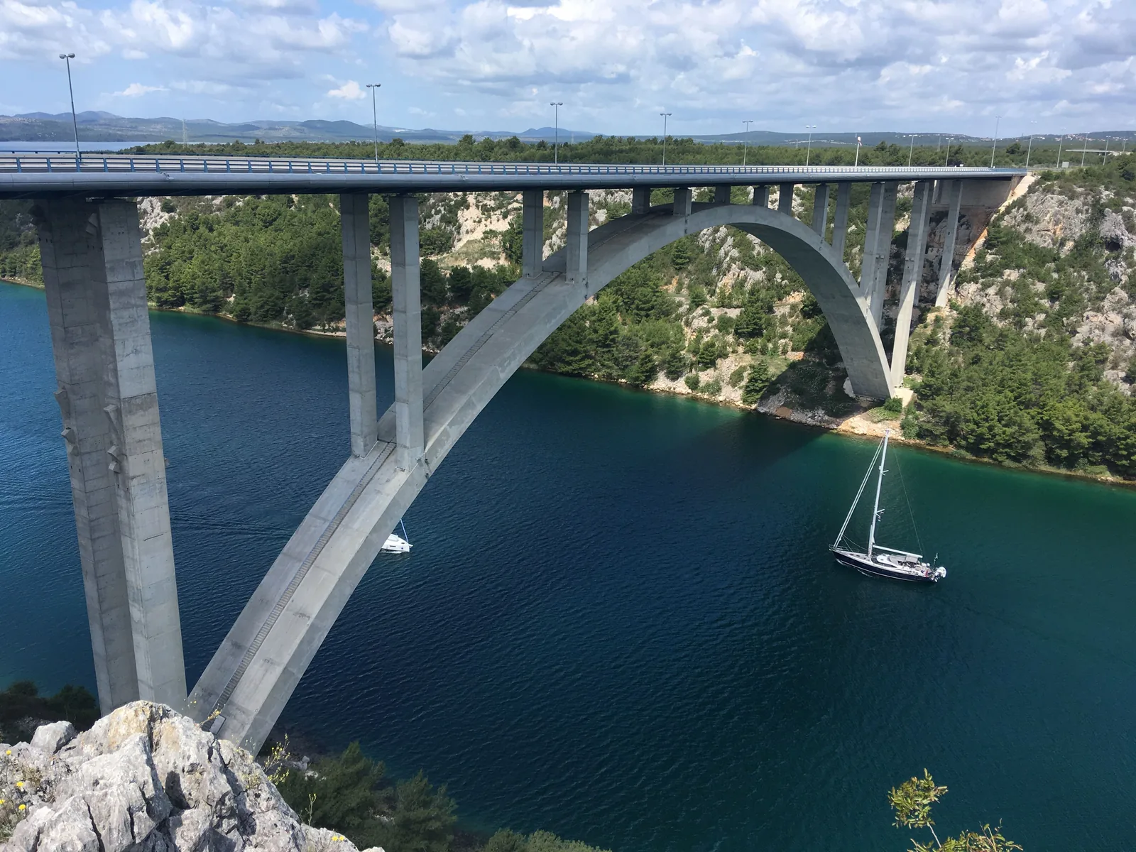 A concrete arch road bridge over a deep river gorge lined with pines and pale limestone, with a sailing yacht on the water below