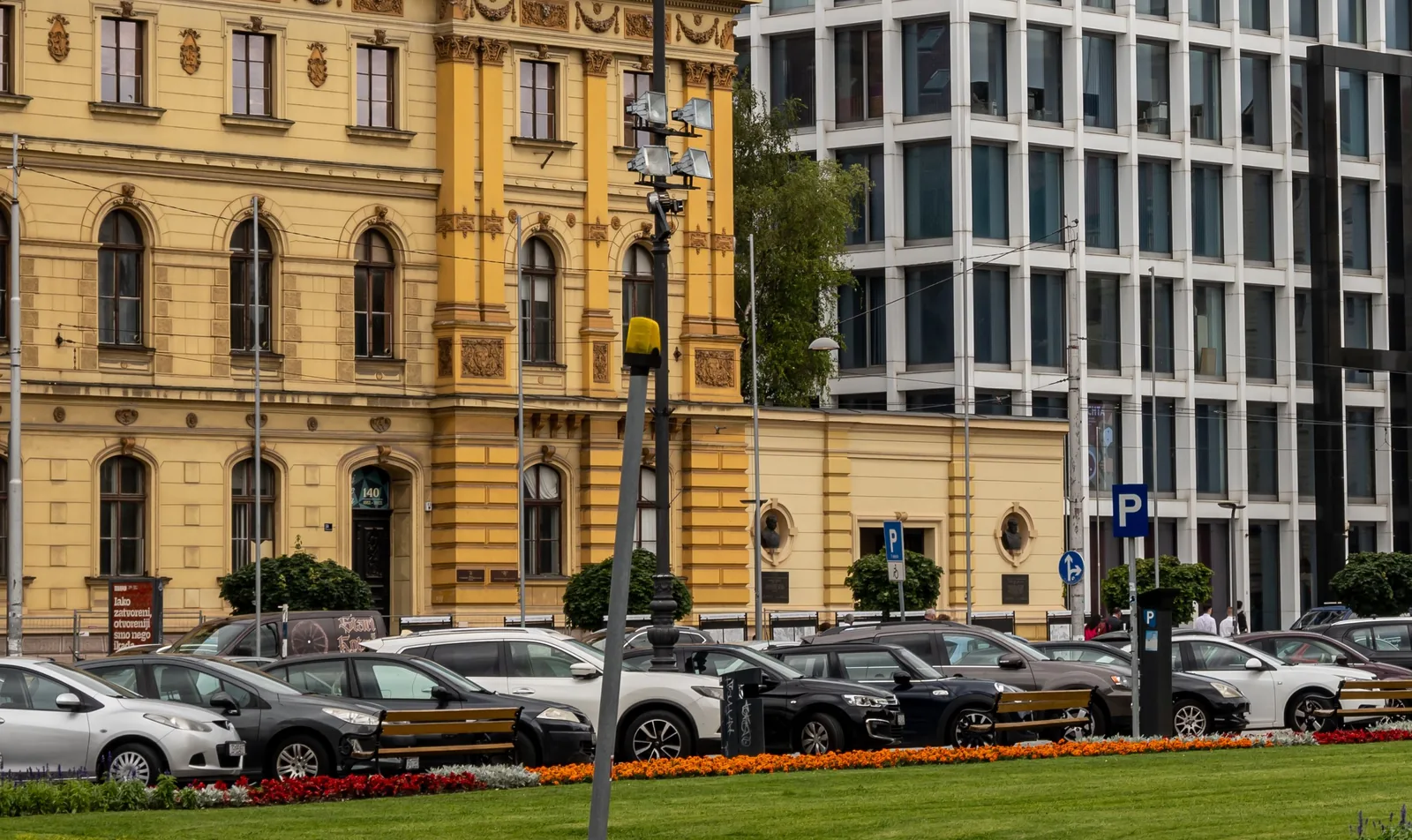 Coches aparcados en fila junto a un parquímetro y señales azules de P, delante de una fachada histórica amarilla en Zagreb
